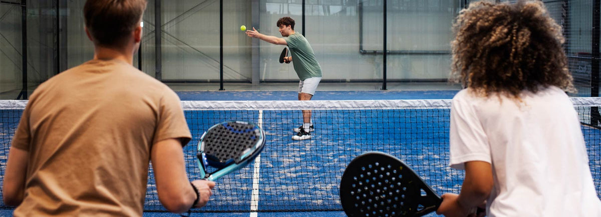 Twee toeschouwers kijken naar een padelwedstrijd op een blauwe baan